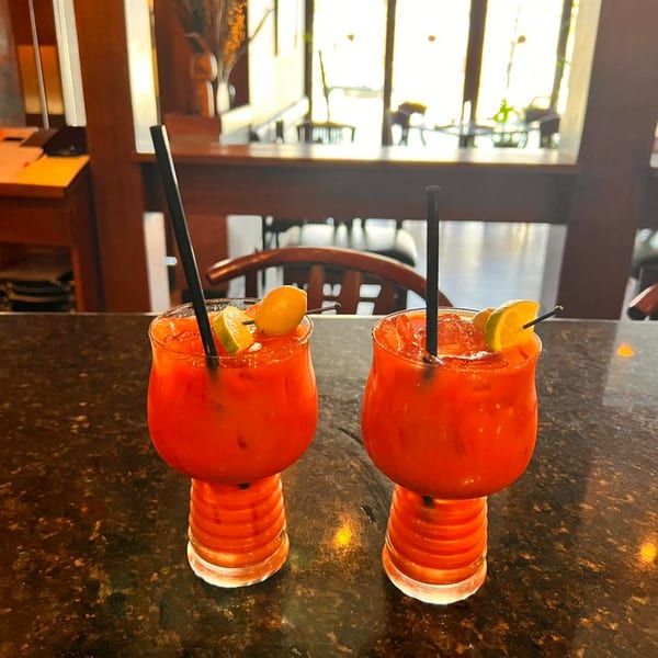 Two vibrant red cocktails with lime and kumquat garnishes on a dark bar, with a bright window in the background.