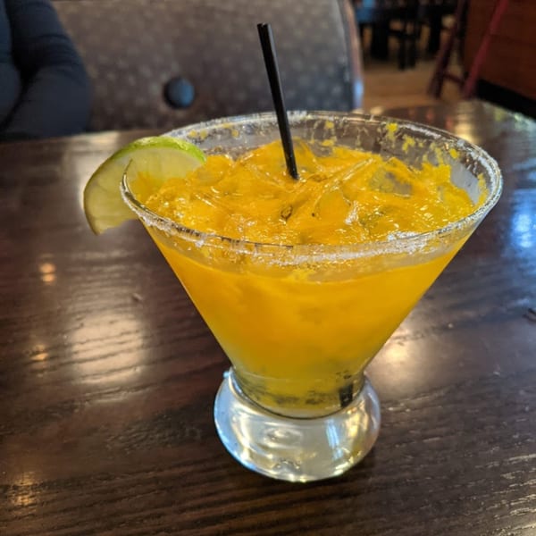A vibrant yellow mango margarita with a salted rim, lime wedge, and black straw on a wooden table.
