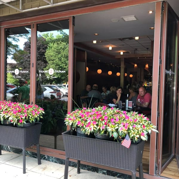 House of Hunan restaurant exterior with large windows, potted pink flowers, and patrons dining inside.